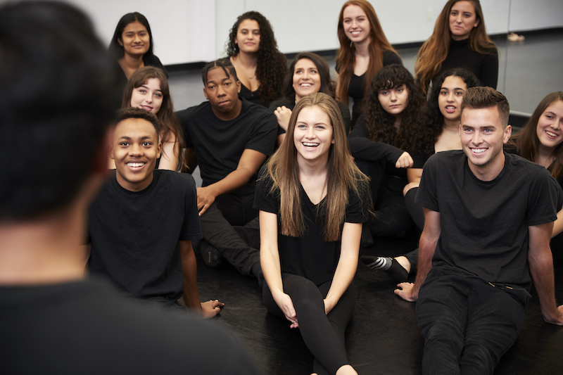 Teacher At Performing Arts School Talking To Students Sitting On Floor In Rehearsal Studio