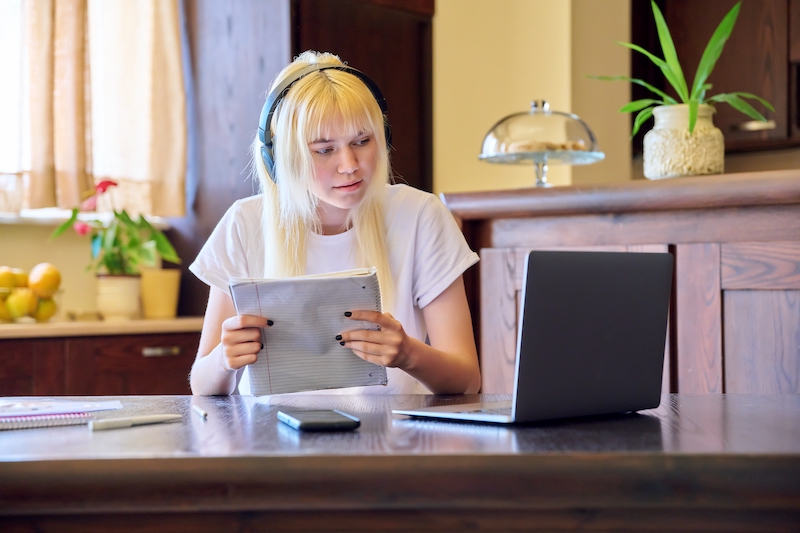 Female student with headphones writing a research paper for school withwith a laptop and notebook 