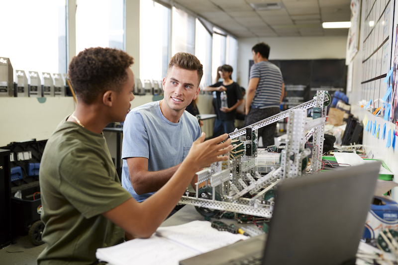 Two engineering students discuss a project in class sitting at a desk with a laptop