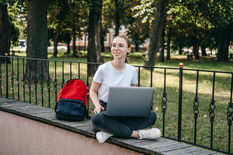 Female student sitting with laptop and backpack outside