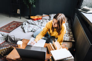 Young woman college student studying with laptop, distantly preparing for test exam, writing essay doing homework at home, distantly education concept.