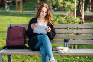 Girl reading a college acceptance letter on a bench outside with her backpack next to her