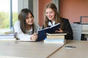 Two fifteen-year-old schoolgirls reading books and taking abstract in copybooks are doing homework in the school library.