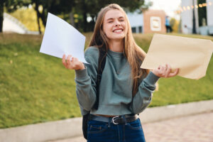 Pretty cheerful casual student girl opening envelope with application results rejoicing in city park