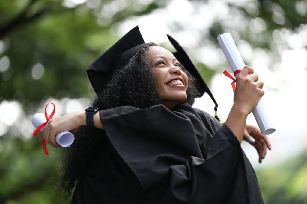 Two graduates in caps and gowns joyfully celebrate, each holding a diploma with smiles and raised hands. Their excitement shines in an outdoor park with greenery, marking a proud moment of achievement