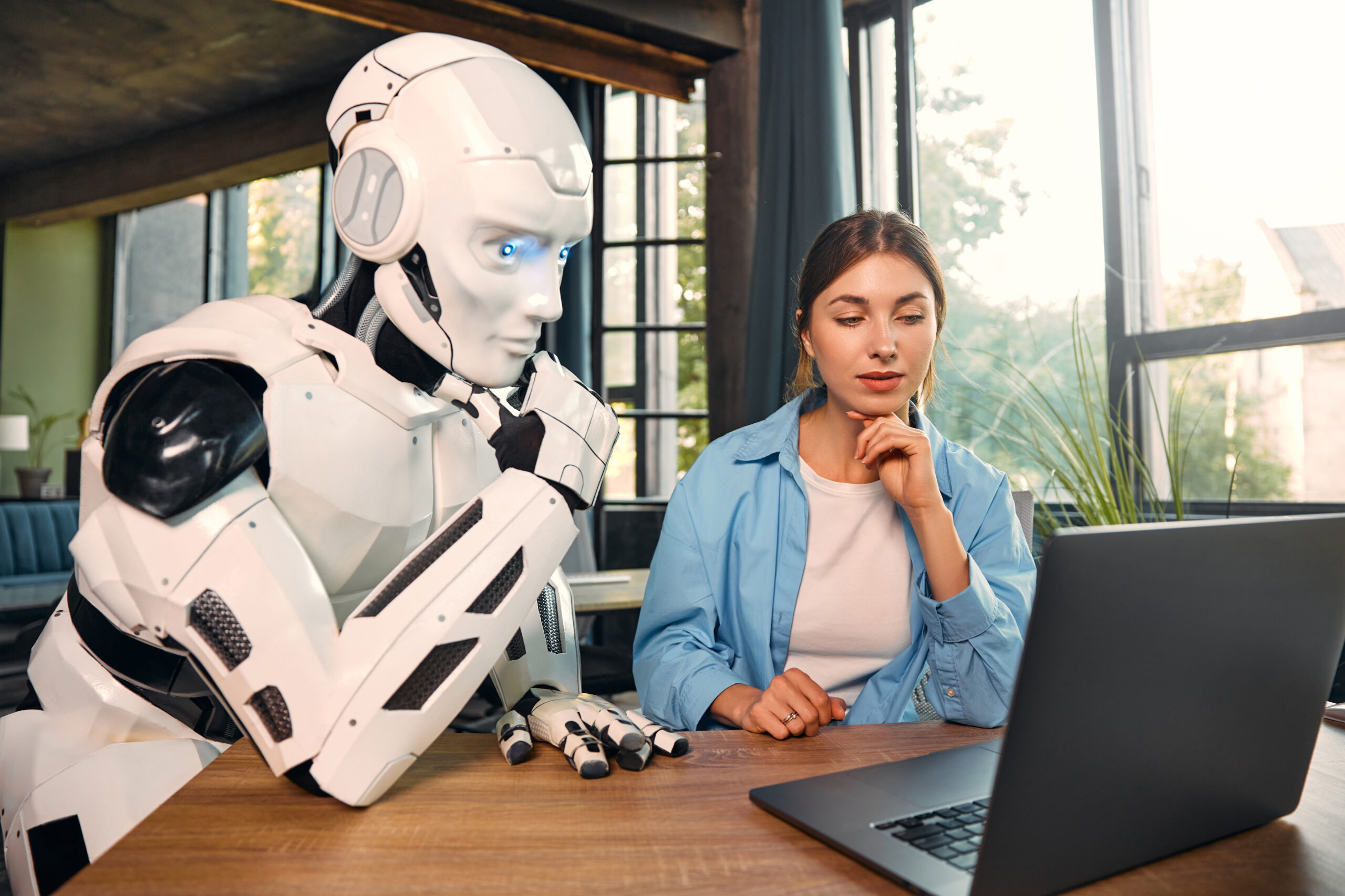 Young woman and a humanoid robot working while sitting at a laptop in a modern office. Collaboration between humans and artificial intelligence.